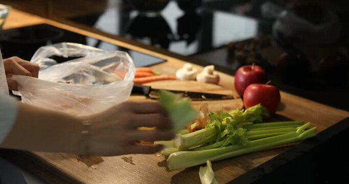 Cropped woman hands move away organic food scrapes waste in plastic bag on wooden table. Zero waste, degradable food. 