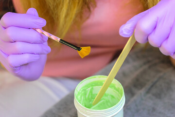 A cosmetologist wearing medical gloves stirs cream in a jar, making a face mask using a makeup brush. Grooming cosmetics for women. Beautician professional master preparing for female beauty procedure
