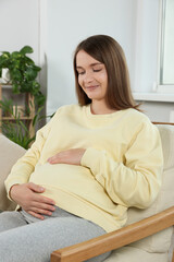 Happy pregnant woman sitting on armchair and touching belly in living room