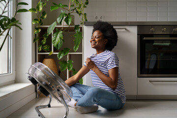 Young happy satisfied African American woman sitting on floor in kitchen in front of air cooler,...