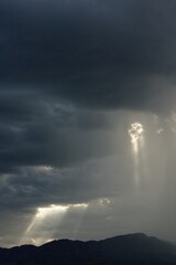 Sunbeams pierce a rain cloud with rain to one side and mountains below the clouds.