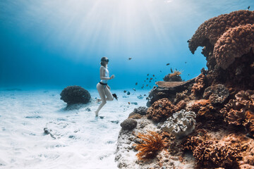 Freediver girl in bikini underwater on sandy bottom near coral reef with tropical fish in...