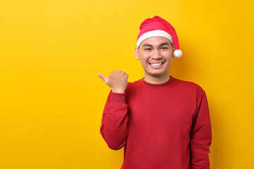 Fototapeta na wymiar Smiling young Asian man in Santa hat pointing thumb aside at blank space for text on yellow studio background. celebration Christmas holiday and New Year concept