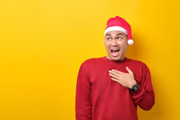 Surprised young Asian man in Santa hat looking aside at copy space on yellow studio background. celebration Christmas holiday and New Year concept