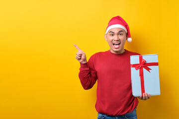 Surprised young Asian man in Santa hat holding gift box with ribbon, pointing finger aside at copy space on yellow studio background. celebration Christmas holiday and New Year concept