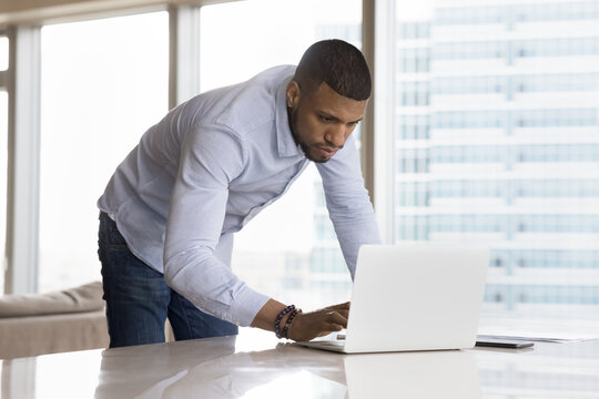 Serious busy handsome African business leader man using online app on laptop, wireless technology for communication, standing at workplace, bending over work table, typing