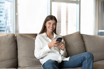 Happy positive young cellphone user woman sitting on home sofa in city apartment, holding mobile phone, using gadget for Internet communication, looking away, smiling