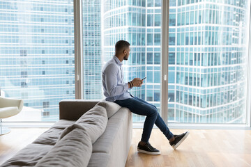 Thoughtful dreamy millennial Black man using smartphone in office lobby, hotel room, at home,...