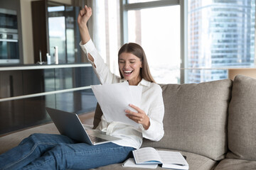 Cheerful young student woman excited with good news, reading paper document at laptop, resting on sofa at home, making winner hand, fist, smiling, celebrating success, laughing