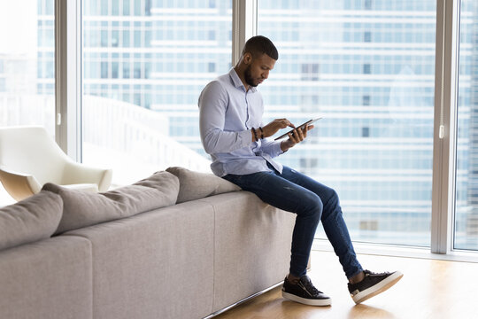 Focused Millennial African Tablet User Man Reading Text On Digital Device Screen, Chatting, Touching Screen, Using Gadget For Online Internet Business Communication, Standing At Home Sofa,