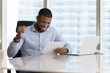 Happy excited young Black businessman getting good news letter, reading paper document at office workplace, making winner yes hand, smiling, laughing, celebrating job success, achieve