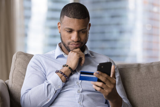 Thoughtful African credit card user holding mobile phone, looking at screen, touching chin in doubts, making decision, using ecommerce banking app for making payment, shopping