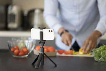 Male cook blogger slicing fresh vegetable ingredients at smartphone webcam, shooting video on cellphone in kitchen, using gadget fixed on tripod, promoting healthy eating, lifestyle. Close up shot
