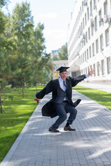 Old happy man in graduation gown jumping outdoors and holding diploma. Vertical.