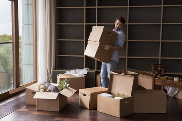 Busy serious renter man carrying stack of paper cardboard relocation boxes in empty room with pile...