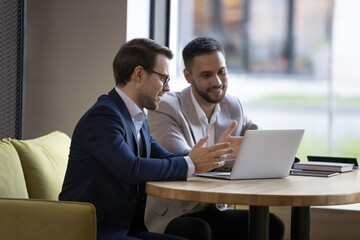 Two young businessmen in formal suits discuss new software sit at table with laptop in workspace, work on startup project, manager offering, presenting new services to client on device meet in office