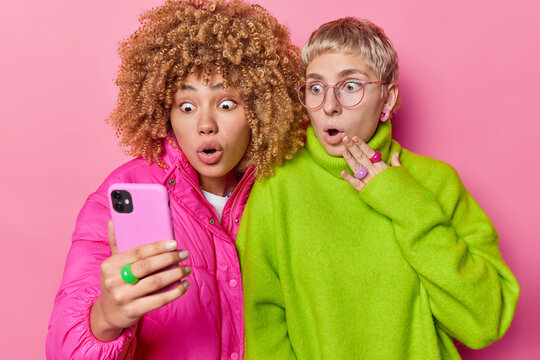 Image of shocked women stand in smartphone screen react to something awful have widely opened eyes wear bright fashionable clothes isolated on pink background. Stupefied female friends with cellular