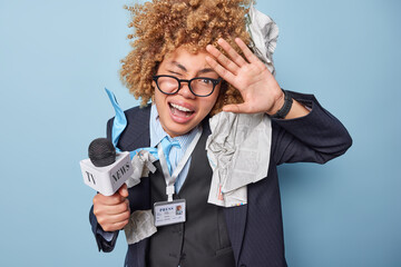 Funny female correspondent keeps palm raised winks eye holds microphone tells breaking news review makes report wears spectacles black formal jacket blue background. Crazy female media worker