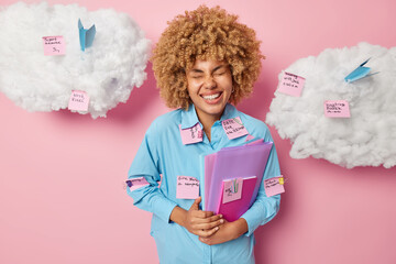 Happy female student stands with folders and memo stickers around laughs with closed eyes glad to finish work in time has busy working week isolated over pink background white clouds around.