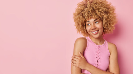 Waist up shot of woman with curly hair dressed in casual t shirt stands bare shoulders smiles gently focused aside dreams about something pleasant isolated on pink background blank space for your text