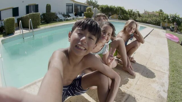 Boy making selfie of himself and his friends at swimming pool on warm summer day. Camera point of view. Children showing tongue and hand gestures, having fun. Modern technologies, friendship concept.
