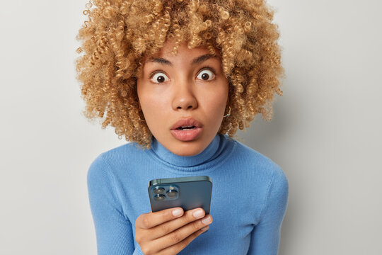 Photo of shocked curly haired woman with amazed expression stares at camera cannot believe own eyes holds smartphone finds out shocking news wears blue turtleneck isolated over grey background