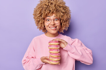 Photo of lovely curly haired woman bites lips holds stack of delicious glazed doughnuts has sweet...