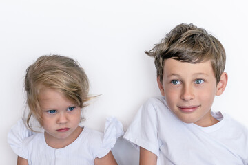Portrait of a brother and sister against a white wall. A boy 9 years old and a girl 2 years old white Caucasian in white clothes stand near the wall during the holiday