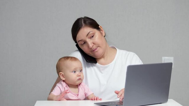 Brunette mother talks with colleague on smartphone and takes care of baby daughter on lap at same time. Baby girl looks with puzzled expression at laptop