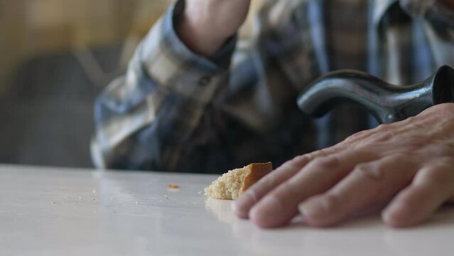 close-up of a pensioner with a walking stick sitting at the table eating a piece of bread. selective focus. high inflation, financial crisis, food shortage, the pensioner is starving