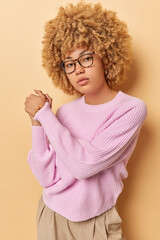 Self confident woman looks seriously at camera keeps hands together has determined assured expression wears eyeglasses long sleeved pink jumper and trousers stands indoors against beige background