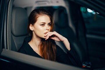 Obraz na płótnie Canvas a stylish, luxurious woman sits in a black car at night, touching her face with a thoughtful expression. Close horizontal photo