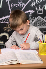 a little boy of Slavic European appearance writes with his left hand at the table against the background of a slate wall