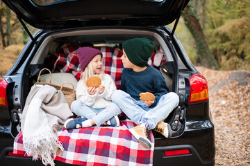 Happy smiling kid girl and boy eating biscuits with kid girl sitting in car body with blanket and decoration outdoor. Childhood.