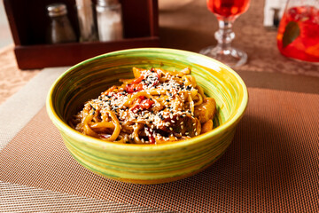 Udon wok stir fry noodles with vegetables, chicken meat and sesame seeds in green bowl on noisy brown table mat background in outdoor restaurant. Front view, lifestyle