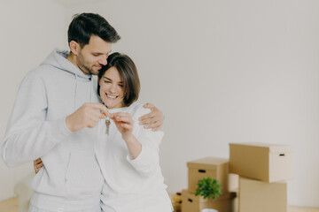 Delighted woman and man home owners enjoy buying new flat, hold keys fromm their own apartment, pose against white wall with unpacked cardboad boxes. Family mortgage, ownership, real estate.