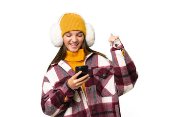 Teenager caucasian girl wearing winter muffs over isolated background with phone in victory position