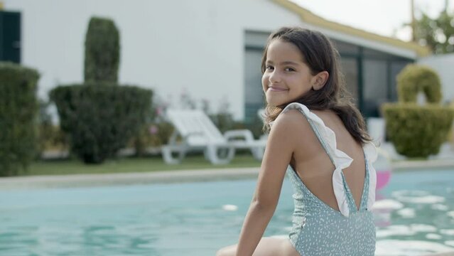 Side view of girl sitting by swimming pool on warm summer day. Young Caucasian girl in swimsuit turning head and smiling to camera. Wind playing with her hair. Childhood, joy concept.