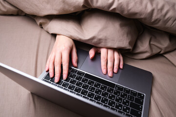 A young woman is working on a laptop in bed wrapped in a blanket. Work from home, freelancer. Focus on hands