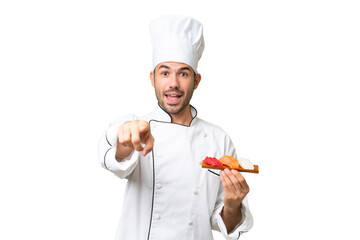 Young caucasian chef holding a sushi over isolated background surprised and pointing front