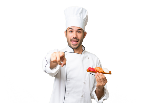 Young caucasian chef holding a sushi over isolated background surprised and pointing front