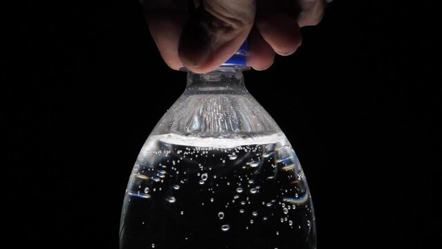A bottle of carbonated water is unscrewed by hand on a black background. Bubbles come out of the bottle. Pure mineralized water with gas. Man opens plastic bottle of carbonated water, black background