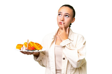 Young Arab woman holding waffles over isolated background having doubts while looking up