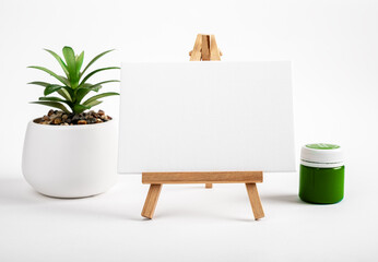Blank white canvas mockup on mini easel and potted house plant, green gouache paint in jar on desk