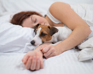 Caucasian red-haired woman sleeps in an embrace with a jack russell terrier dog on a white sheet.