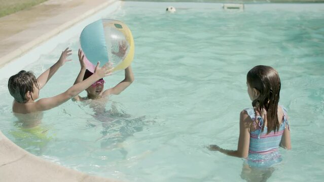 Children playing with inflatable ball in swimming pool outside. Group of boys and girls having fun, throwing ball to each other while standing in water. Childhood, summer, joy concept.