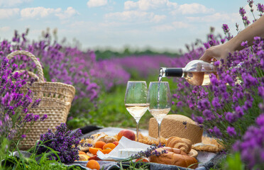 Wicker basket with delicious food for a romantic picnic in a lavender field. The girl pours wine.