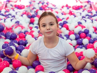 Fototapeta na wymiar A little girl is sitting in a large dry children's pool with colorful plastic balls in the game center looking into the frame holding balls in her hands. Children's entertainment