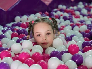 Fototapeta na wymiar A little surprised girl lying on colorful plastic balls in a large dry children's pool in the play center. Smiling at the camera. Close-up portrait. Birthday fun