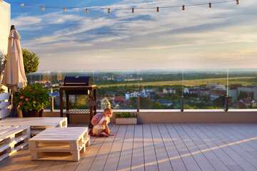 cute baby girl sitting on sunlit rooftop patio at summer evening with beautiful landscape view